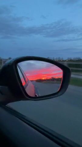 The view from a car's side mirror shows a vivid sunset over a road. The sky is a mix of deep blue and vibrant red-orange hues, reflected in the mirror. Raindrops cling to the mirror's surface. The road stretches into the distance, flanked by fields and sparse vegetation. The car is moving, as evidenced by the blurred background and the changing view in the mirror.
