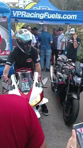 A motorcycle rider wearing a black t-shirt, blue jeans, black sneakers, a colorful helmet, and gloves sits on a red and white motorcycle. The motorcycle has the number 058 on its front. Another black motorcycle is parked to its right. Behind the rider, people stand around under a tent with "VPRacingFuels.com" printed on it. The area is paved. A person in a red shirt stands in the foreground, partially blocking the view. Several people are observing the rider and motorcycle.