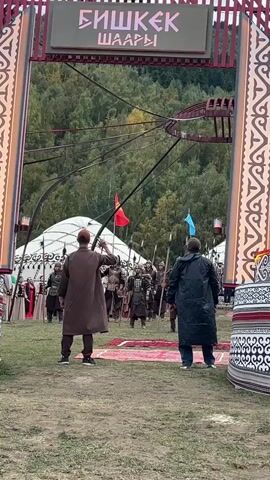The video shows an outdoor scene with two men in the foreground facing away from the camera. They are standing on a grassy area, and beyond them is an arched gateway with signage in Cyrillic at the top. Behind the gateway, a group of people dressed in historical or fantasy-themed costumes, resembling warriors with armor and spears, are approaching. Some of them are holding flags. There are tents in the background, and a forested hillside is visible beyond that. The overall impression is that of a staged event or reenactment.