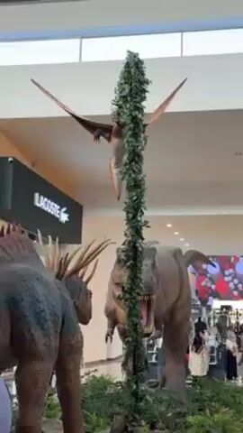 The scene captures a static display of dinosaur models inside a shopping mall. In the foreground, a Spinosaurus model is positioned with its distinctive sail-like structure. Behind it, a Tyrannosaurus Rex model stands with its mouth open, displaying its teeth. Between the two dinosaurs, a tall pole wrapped in artificial foliage stands. At the top of the pole, a Pterodactyl model is suspended. In the background, the recognizable logo for the clothing brand Lacoste is visible, along with a large advertisement with red and white imagery. Several mall patrons stand near the display, casually observing and taking pictures. The mall environment is well-lit, with a high ceiling and a modern architectural design.