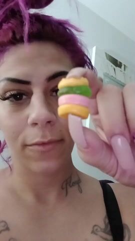 A woman with purple hair is holding a small gummy candy in the shape of a burger, made with yellow, green, and pink layers. She brings the candy to her mouth and eats it. She smiles slightly. The woman has multiple tattoos on her chest and neck. There is a piece of artwork on the wall behind her.