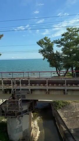 The video captures a view from a moving train, looking out towards a body of water, likely the sea or a large lake. In the foreground, train tracks are visible, indicating the perspective of the viewer. Beyond the tracks, there is a concrete structure, possibly a bridge abutment or retaining wall, with a small channel of water running beneath it. Further beyond this, a raised railway bridge is seen with a metal railing. To the right of the bridge, there are trees with green foliage. The water is a vibrant blue, meeting a clear sky with scattered white clouds. Along the shoreline, several buildings with red roofs are visible, along with some boats. There are also utility poles with electrical wires stretching across the frame. The scene is bright and appears to be under daylight.