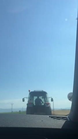 The video shows the rear view of a tractor on a sunny day. The tractor is dark gray and blue with a light on top. A person is visible in the driver's seat. The tractor is on a road with a clear blue sky in the background. There are some telephone poles and a few faint lines of trees or buildings in the distance. The video is taken from inside a vehicle, as evidenced by the dashboard in the foreground and the edge of the windshield on the right. There is a small speck on the windshield. The tractor is stationary at the beginning of the clip and then begins to move forward.