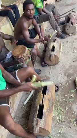 The video shows a group of men and a young boy engaged in a musical performance or practice in an outdoor setting in Unwana, Nigeria. Most participants are seated on concrete steps.

In the foreground and middle ground, several men are actively playing traditional instruments. Two men are striking large, hollowed-out wooden logs (resembling slit drums) with sticks that appear to be segments of sugarcane or similar plant material, creating rhythmic sounds. One man, shirtless, plays a djembe drum with his hands. The performers are focused on their instruments, with their bodies moving to the rhythm. The man playing the djembe and another man playing one of the log drums look directly at the camera and smile, indicating awareness and a friendly demeanor.

Other men and a boy are seated around, observing or participating. One man in a green tank top with white text (partially visible) is also playing a log drum. Another man wears a green polo shirt, and a young boy in a plaid shirt sits in the background. The general attire is casual, with some men shirtless or in simple shirts. The setting appears to be a community space, possibly a courtyard or a village gathering area, with a dusty ground surface and concrete seating. The activity takes place during the daytime, under bright natural light. No discernible text on objects provides additional context beyond standard branding.