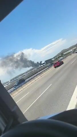 The video captures a view from inside a vehicle, likely a truck, looking out through the windshield at a highway. A plume of dark smoke is visible in the distance on the left side of the frame, originating from what appears to be a vehicle fire or industrial incident. Several other vehicles, including trucks and cars, are on the highway, moving in the same direction as the camera's perspective. The sky is blue with some white clouds, and the landscape in the background suggests a semi-urban or industrial area with some hills. The camera angle is slightly tilted, giving a dynamic perspective of the scene.