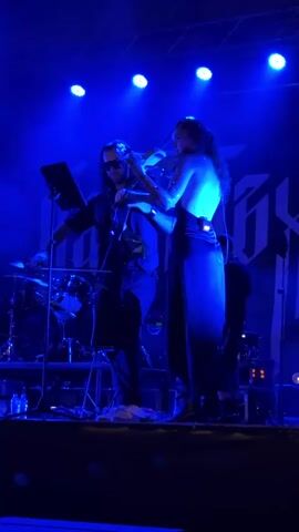 The media file shows a live musical performance on a stage bathed in vibrant blue light. Two individuals are prominently featured. On the left, a person wearing sunglasses and a dark outfit is playing a cello. They are positioned behind a music stand displaying a tablet. To the right, a woman in a shimmering, sleeveless blue gown is playing the violin. Her back is to the camera, highlighting the open back of her dress. Behind them, a drum set is visible, along with other stage equipment and lighting. The overall atmosphere is dark and moody, typical of a concert setting. The bright blue stage lighting creates a dramatic effect, illuminating the performers and their instruments. The backdrop features a large, stylized logo or graphic in a lighter shade against the darker stage. The performance appears to be taking place indoors, likely in a venue in Sofia, Bulgaria, given the location context. The focus on the musicians and the energetic lighting suggest an intense and captivating performance.