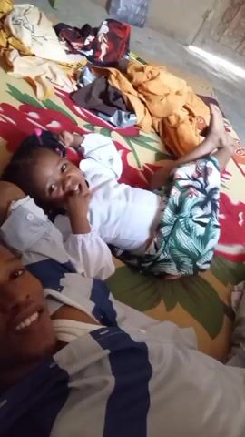 A man and a young girl lie on a bed with a colorful floral pattern. The man is in the lower left, smiling and taking the video. He is wearing a striped shirt. The girl is lying on her back next to him, looking at the camera with a finger in her mouth. She is wearing a white top and floral patterned bottoms. Several articles of clothing are scattered on the bed around them in shades of yellow, orange, red, and brown. The room appears to be indoors and well-lit.