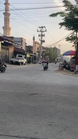 A street scene with a mosque in the background. Several motorcycles and a small truck are visible on the road. A woman is riding a bicycle with two baskets attached. The sky is clear and the sun is low, suggesting it is either early morning or late afternoon.