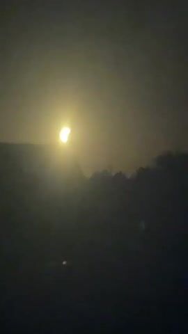 This is a short, silent video of a partial solar eclipse. The sun is visible in the upper portion of the frame, partially obscured by the moon. The obscured portion of the sun has a crescent shape. The sun is bright and yellow, with some hazy clouds or atmosphere obscuring the view. The lower portion of the frame is dark and appears to be landscape, possibly mountains or hills, also covered in a haze. The video is taken from a distance, and the focus is on the sun and the eclipse. The overall atmosphere is somewhat muted due to the haze and the nature of the eclipse.