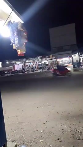 This is a low-light, handheld video captured at night, seemingly from a street-level perspective. In the foreground, a dirt or gravel surface is visible with some scattered debris. To the left and slightly out of focus, a portion of a blue structure is present. Above and to the left, hanging from an overhang, are various packaged goods, possibly snacks or small items, illuminated by a bright light. The background depicts a row of shops or stalls, many of which are lit, suggesting they are open for business. Several motorcycles and scooters are parked in front of the establishments or are in motion on the street, indicated by streaks of light and blurred shapes. Some figures, presumably people, are visible walking or standing in front of the shops. A sign with text is visible on one of the stalls, but it is illegible due to the distance and lighting. The overall impression is of a bustling night market or commercial area.