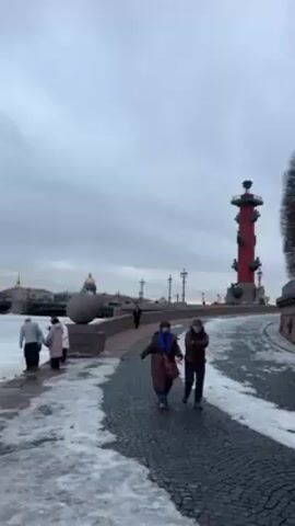 The video shows a winter scene in Saint Petersburg, Russia. The sky is overcast, and snow and ice are visible. In the foreground, two people walk along a cobblestone path, past a Rostral Column. Other people are on the left edge of the frame, near the Neva River's edge. Some people are throwing objects, possibly stones or ice chunks, into the frozen river. Buildings and landmarks are visible across the river in the background, including the Winter Palace, Admiralty Building, and St. Isaac's Cathedral.