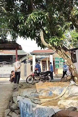 A group of people are gathered outside a building. A red motorcycle is parked in the foreground. There is a tree with green leaves providing shade. The ground is covered with rocks and a tarp. The people appear to be in casual conversation. Some are wearing blue shirts. One person is wearing a red and white outfit. Another person is squatting on the ground.
