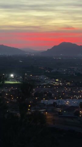 The video is a static, elevated shot of a town at dusk. Above the town, the sky transitions from gray to yellow to a band of red. In the distance, silhouetted mountains can be seen. The town itself is dark, but lit by numerous, regularly spaced orange streetlights. There are some trees in the foreground. In the audio, there's the sound of children's voices.