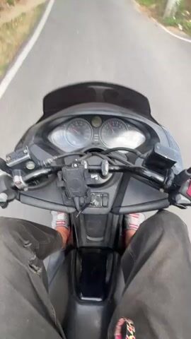 The video shows a first-person perspective of someone riding a black scooter on a paved road lined with trees. The rider is wearing grey jeans and casual shoes with colorful patterns. The scooter has a dashboard with gauges and controls, and a smartphone mount. The road curves gently ahead, and other scooters can be seen in the distance, also traveling in the same direction. The environment is green with lush trees on either side of the road, and utility poles are visible along the roadside. The sky is blue with some clouds.