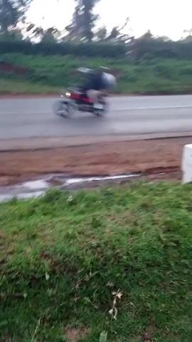 The video starts with a blurred view of a motorcycle speeding on a gray road with green and red-brown land beside it. The camera then abruptly moves, blurring the view of the road and surrounding land. Subsequently, a person wearing blue jeans and light-colored shoes walks into the frame. The camera follows them as they walk along the green and red-brown land. The video concludes with a focused view of the red-brown soil.