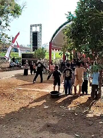 A group of people are gathered outdoors around a dirt area. In the center, two individuals are engaged in a demonstration. One person, dressed in black with a white bandana on their head, wields a curved stick or whip. The other person, wearing a striped shirt and a sarong-like garment, is positioned opposite them. A cloud of dust or powder is visible in the air between them, suggesting some kind of impact or spray. Several spectators, including children and adults, watch the event. In the background, large speakers are set up, and a tent with red and white draping provides shade. The environment appears to be rural, with greenery visible beyond the immediate gathering.