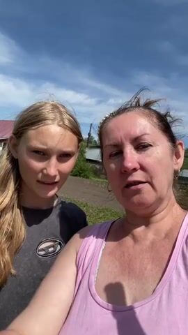 A close-up selfie-style video shows two females outdoors on a sunny day. The camera is held by an adult woman in the foreground, wearing a pink tank top. She has brown hair pulled back, and her face is lightly tanned. She gestures with her right hand, holding it up as if waving, and speaks cheerfully into the camera. Beside her, a younger girl with long, blonde hair and a dark t-shirt smiles subtly at the camera. The background features a rural or suburban landscape under a bright blue sky with some white clouds. Several houses with red or blue roofs are visible in the distance, surrounded by green grass and what appears to be tilled earth or garden beds. The overall mood is positive and casual.