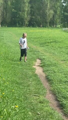 A boy is walking along a dirt path surrounded by green grass in a park-like setting. He is wearing a white t-shirt with a graphic, black cargo shorts, and Crocs. He is holding a water bottle in his left hand. The grass is dotted with yellow flowers, and there are trees in the background. The boy is walking towards the camera.
