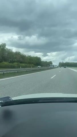 A view from inside a car, of a multilane highway. On the highway are numerous cars and trucks, including one large truck carrying a wind turbine blade. The sky is overcast. The highway has trees alongside it. The camera is stationary.