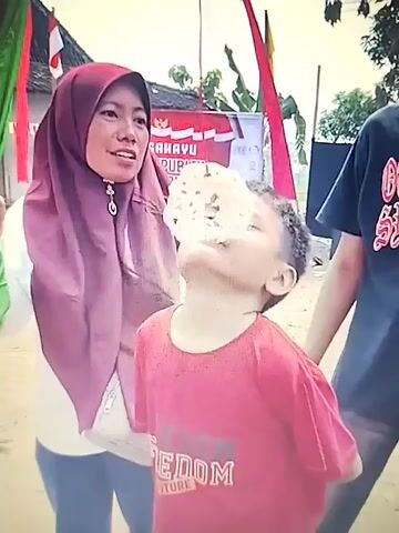 A woman wearing a maroon hijab and white shirt stands to the left of a young boy in a red t-shirt. The woman holds a white, circular object on a string in front of the boy. The boy is looking up at the object and appears to be trying to bite it. Another person's arm is visible on the right side of the frame, holding the boy. In the background, there is a banner with Indonesian text and flags. The scene appears to be an outdoor event.