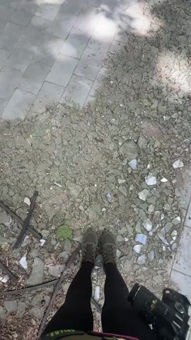 A person stands on a ground littered with debris, including shattered glass. The camera slowly pans up to reveal an abandoned indoor swimming pool. The pool is empty and dilapidated, with cracked tiles and overgrown vegetation visible through large windows. The roof is rusted and partially collapsed.