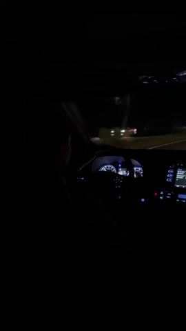 A person is driving a car at night. The dashboard is illuminated with blue lights. The car is moving on a road with a dividing line. The exterior environment is dark with minimal lighting.