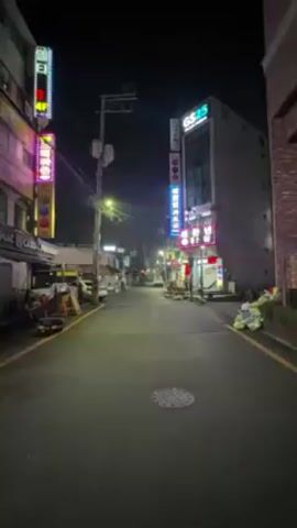 A blonde woman walks down a narrow street at night. She is wearing a black leather jacket and a baseball cap. The street is lined with buildings with neon signs.