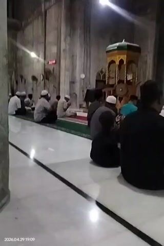 This video captures an indoor scene within a mosque or Islamic prayer hall. Several men and a child are seated on the floor, some on prayer mats, while others sit directly on light-colored tiled flooring. Most of the men are wearing traditional Muslim caps (kufis or peci). Many have their hands raised in supplication or prayer (dua). In the background, a decorative mihrab or minbar structure, colored yellow and green, is visible. The walls appear to be made of stone or concrete. A bright light source is overhead, creating reflections on the floor. A red digital clock display is on the wall, though the numbers are not clearly legible. The video's timestamp indicates the time as 7:09 PM on April 29, 2026, consistent with an evening religious gathering. A male voice reciting Arabic text is audible in the background.