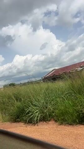 This media file captures a scenic view of a rural landscape in Jakatai, Nigeria. The scene is dominated by lush green grasses and vegetation, with a dirt road in the foreground. In the middle ground, several buildings with corrugated iron roofs are visible, interspersed with trees. The sky is filled with dramatic, voluminous clouds, suggesting a dynamic weather pattern, possibly a prelude to rain or the aftermath of a storm. The lighting indicates it is daytime, likely late morning or early afternoon. The overall atmosphere is serene and natural, showcasing a typical rural setting. There are no people or explicit objects of human activity beyond the buildings, which appear to be houses or small structures. The activity implied is the passage of time and the natural processes of the environment.