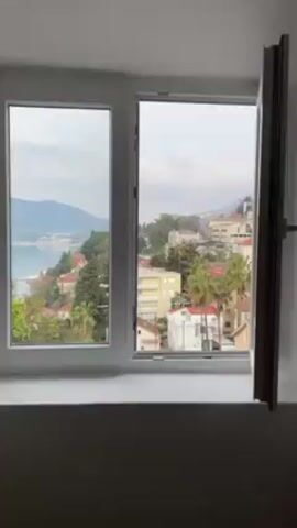 The video starts inside, looking out a window. The window is white and has two panes. One pane is open, revealing a view of a town on a hill, with a body of water in the distance. The town has many buildings with red-tiled roofs. There are also some palm trees and other green vegetation scattered throughout. The sky is overcast and the light is somewhat muted. The camera zooms in slowly on the view outside the window, focusing on the rooftops, trees and buildings of the town. There are no people visible in the video.
