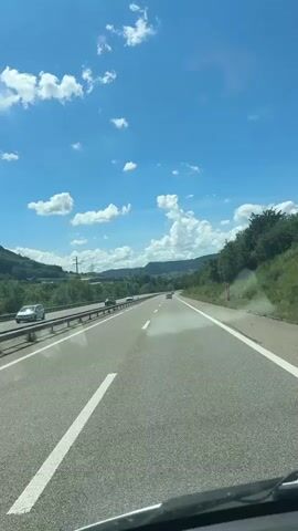 The video shows a car driving on a highway on a sunny day. The sky is blue with scattered white clouds. The road has two lanes in each direction, separated by a median. There are a few other cars on the road. The surrounding landscape is green and hilly. The video is shot from the perspective of the driver.