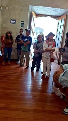 The video shows a museum tour group standing in a room with wood floors. A woman in a white jacket and black pants, with a yellow tote bag, is speaking to the group.  She's gesturing towards two large portraits in gold frames hanging on a wall. The walls are light-colored, with a sign in Spanish visible near the ceiling. Some of the group members are holding books or papers.  An open doorway is visible in the background, leading to a brighter outdoor area. The speaker appears to be explaining the paintings to the group.
