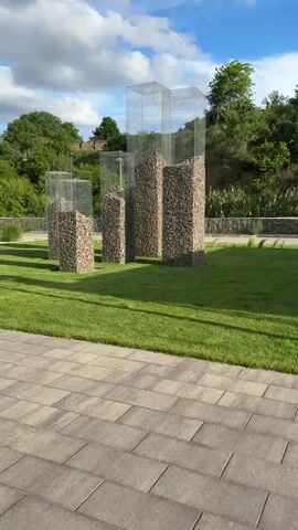 The video presents an outdoor scene featuring several modern art installations under a clear, bright blue sky with scattered white clouds. In the immediate foreground, a paved walkway made of rectangular grey pavers runs horizontally across the bottom of the frame.

Beyond the walkway, a well-maintained green lawn serves as the setting for five distinct, tall, rectangular sculptures. Each sculpture is composed of two main parts: the lower section consists of a gabion, a wire cage filled with small, light-colored stones or rocks, forming a solid base. Extending upwards from these rock-filled bases are empty wire cage structures, creating an abstract, skeletal framework that suggests the form of unfinished buildings or a modern urban skyline. The sculptures vary in height, with some appearing significantly taller than others, creating a dynamic visual composition.

In the background, a dense line of lush green trees and foliage provides a natural backdrop, suggesting a park or a landscaped public area. The sunlight is bright, indicating either late morning or early afternoon. The weather appears pleasant and clear. No people are visible within the frame. The audio captures the distinct chirping of birds and faint, distant human voices, contributing to a serene outdoor ambiance. No text is visible in the video.