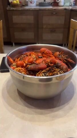 The video shows a close-up, slightly elevated, static shot of a large metal bowl filled with cooked crawfish. The crawfish are primarily reddish-orange, with some appearing darker. They are piled high in the bowl, obscuring the bottom. The bowl is placed on a light-colored, speckled countertop. In the background, out of focus, are kitchen cabinets with wooden doors and a portion of a chair. There is a black object, possibly a smartphone, on the countertop to the left of the bowl. The lighting is warm and appears to be coming from overhead. There are no people visible in the video.