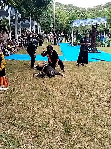 The video shows an outdoor performance on a grassy field, with spectators seated under white tents. In the foreground, a person is lying on the ground while being attended to by two other people, one kneeling beside them. A fourth individual in a striped shirt and traditional attire stands nearby, observing. A man in black holds a camera or phone. In the background, more people are standing around, seemingly watching the performance. There is some foliage and a tall structure behind the crowd.