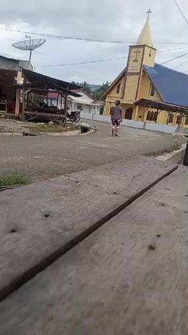 The video shows an outdoor scene with a yellow church with a blue roof in the background. A person wearing a checkered shirt and shorts is walking away from the camera on a paved road. To the left of the road, there is a small building with a satellite dish on top. A motorcycle is parked near the building. The foreground of the video is dominated by a close-up view of weathered wooden planks. The sky is overcast.