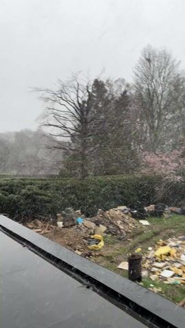 The video shows an outdoor scene on a snowy day. The camera appears to be positioned inside, looking out through a window. Snowflakes are falling heavily. In the foreground, the edge of the window frame is visible. Beyond the window, there is a garden or yard covered in green grass. Scattered throughout the yard are various debris and discarded materials including yellow insulation, pieces of wood, and other construction-related waste. A green hedge is present in the middle ground, with bare trees and some greenery in the background. The sky is overcast. There is a sense of untidiness or ongoing construction in the outdoor space.