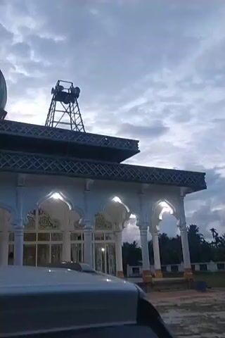 The video shows a mosque under a cloudy sky. The mosque is white with arched windows and a blue and gold dome. A speaker tower is also visible on the roof. A car is parked in front of the mosque. An Islamic call to prayer is playing.