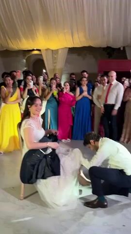 In a video taken from a low angle, a wedding scene unfolds indoors under white draped ceilings, brightly lit. A bride, seated on a wooden chair, dressed in a white gown and veil, looks down and laughs as a groom kneels before her, his head close to her feet. He wears a white shirt, dark pants, and brown shoes. The bride holds a dark jacket on her lap. Surrounding them is a crowd of wedding guests, many holding up phones to capture the moment. Women in vibrant dresses – yellow, teal, hot pink, and royal blue – stand out, as do men in suits or shirts with ties. A photographer with a professional camera is also present. The floor has a patterned design. The atmosphere is celebratory, with laughter and the sound of music audible.