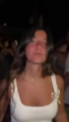 The video, shot at night, appears to be of a celebration or party. The camera begins focused on a young woman with long brown hair and a white top. She is smiling and appears to be dancing. A man in a white shirt is visible behind her. The camera then pans to the right, showing a crowd of people, and the surrounding environment, a street lined with white buildings and streetlights. People are mingling, clapping, and cheering. The camera continues to move, capturing more of the crowd and the setting.