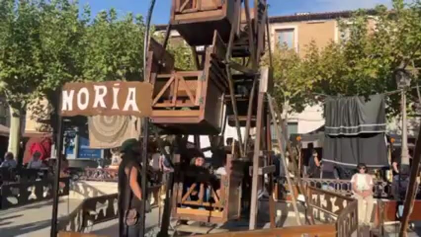 A small, wooden Ferris wheel spins slowly in an outdoor plaza. The Ferris wheel has several enclosed wooden carriages. A woman rides in one of the carriages. A sign next to the ride reads "NORIA". A man stands near the ride, appearing to operate it. People sit at tables behind a short, decorative wooden fence. Trees and buildings are visible in the background under a bright, sunny sky.