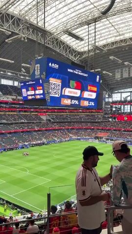 The video shows a wide shot of a soccer stadium from the perspective of a spectator in the stands. The focus is on a large, high-definition video scoreboard that displays a poll asking "WHO WILL WIN THE MATCH?". Two national flags, Portugal and Spain, are shown, with percentages indicating the audience's predictions for each team. The percentages fluctuate throughout the video, starting with Spain slightly ahead and then shifting to Portugal taking the lead. The scoreboard also features a QR code for "PREDICT NOW". Below the scoreboard, the green soccer field is visible with players gathered in a huddle, and a large crowd of spectators fills the stands. In the foreground, two men are looking at their phones, likely participating in the poll. The stadium appears to be modern and spacious, with a high ceiling structure and lighting. The overall atmosphere is that of a live sporting event.