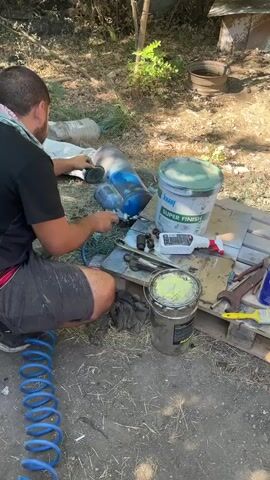A man is kneeling on the ground, wearing a black t-shirt, grey shorts, and a bandana on his head. He is holding a spray gun and appears to be painting a large blue cylindrical object. The object is lying on its side next to him. In the background, there are trees, a large plastic bottle, and a dog house. There are also several buckets and tools scattered around the area, including a large green bucket with "Knauf SUPER FINISH" written on it, a metal bucket with yellow paint, wrenches, and other miscellaneous items. The ground is covered in dirt and straw. The man is actively engaged in the painting process, with the spray gun pointed at the blue cylinder.