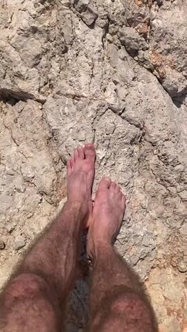 A person's bare feet stand on a large, textured, light-colored rock formation. The camera is positioned above and behind the person, looking down at their feet and the rock. In the background, a dark, pebbly beach meets the sparkling, clear water of the sea, with small waves gently washing ashore. The scene appears to be outdoors on a sunny day.