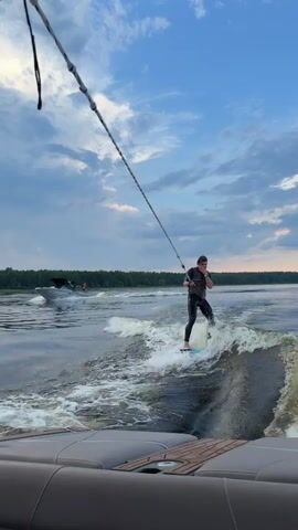 A person is wakesurfing behind a boat on a lake. The person is wearing a wetsuit and holding a tow rope. The boat is a wake boat, which is designed to create a large wake for wakesurfing. The lake is surrounded by trees and the sky is cloudy. The video appears to be filmed from another boat, with a portion of the boat's interior visible in the foreground.