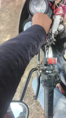 The video begins with a close-up shot of a person's hand holding a motorcycle key. The hand is positioned near the motorcycle's instrument cluster. The person inserts the key into the ignition. The key is then turned, and the instrument cluster lights up, indicating the motorcycle is being started. The camera angle provides a view from the rider's perspective, focusing on the handlebars and the area around the ignition. The background appears to be outdoors, with dirt or gravel visible on the ground. The lighting suggests it is daytime.