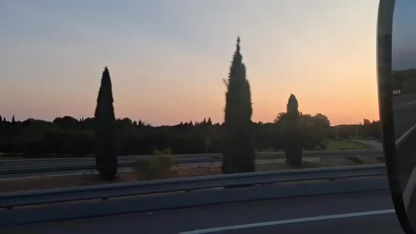 This is a video taken from a vehicle traveling on a highway. The time of day appears to be sunset or sunrise, with the sun visible on the horizon and casting a warm glow across the sky. The landscape includes trees, fields, and a highway with a metal guardrail. The vehicle is moving, indicated by the blur of the scenery and the changing perspective. There are other vehicles visible on the highway in the distance. The video captures a sense of motion and the beauty of the natural light.