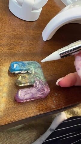 A hand with long, pink nails applies a white liquid with a pen-like applicator to a colorful resin letter “E” on a brown wooden surface. A white gooseneck lamp is visible in the background. The hand is wearing a long-sleeved shirt with black and white stripes, and a sock is visible on the floor.