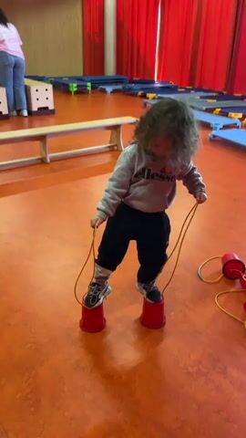 The video features a toddler-aged child with curly dark hair, dressed in a grey hooded sweatshirt displaying the "ellesse" logo and black trousers. The setting is an indoor gymnasium or large play area characterized by a vibrant orange floor. In the background, a light wooden bench, various blue and grey floor mats, and red curtains are visible, suggesting a facility designed for children's activities.

The child is engaged in an activity using two red plastic cup stilts, each with a yellow rope for holding. Initially, the child stands on the stilts, smiling cheerfully at the camera. As the video progresses, the child loses balance and tumbles forward, falling onto the floor while still gripping the ropes attached to the stilts. An adult's voice can be heard reacting with an exclamation, and the child playfully exclaims in French, "Regarde ce que j'ai fait!" (Look what I did!). The child then sits up, seemingly unfazed by the fall, and begins to examine the stilts. The scene conveys a moment of lighthearted play and discovery.