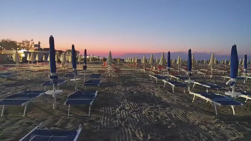 The video shows an aerial view of a beach with rows of empty lounge chairs and umbrellas. The sun is setting, casting a warm glow over the scene. The sky is a gradient of orange, pink, and purple. The sand is dark and appears wet, suggesting it is near the water. There are no people visible in the frame. The overall mood is peaceful and serene.